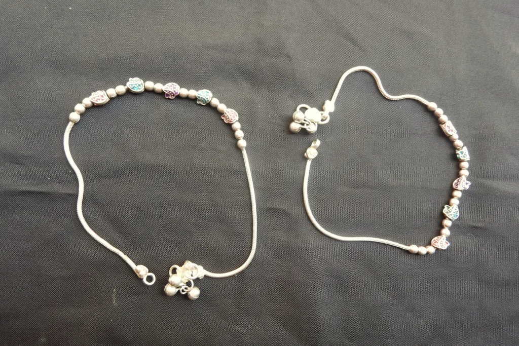 Two traditional Indian silver chain anklets with small bells and coloured beads laid on dark cloth